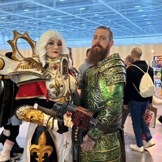 Eine verkleidete Frau und ein Mann in aufwendigen Kostümen schauen auf der Comic Con Stuttgart in die Kamera. 