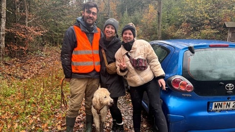 Hannes Weller, Alina Knödler und Anika Siegle schauen fröhlich in die Kamera. Sie stehen hinter Hund Benno und neben einem Auto im Wald.