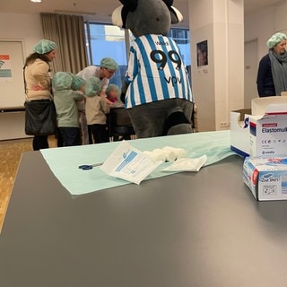 Auf einem Tisch liegt ein Verband. Im Hintergrund malt ein Arzt im Olgahospital in Stuttgart mehreren Kindern eine Wunde auf den Arm. Mit dabei ist auch "Waldi", das Maskottchen der Stuttgarter Kickers. 