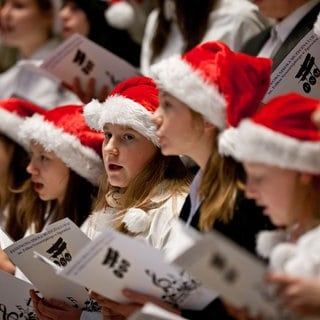 Kinder mit Weihnachtsmann-Mützen singen Weihnachtslieder. Ob in der Kirche, im festlich geschmückten Wohnzimmer oder auf dem Weihnachtsmarkt: Alle Jahre wieder erklingen Weihnachtslieder. 