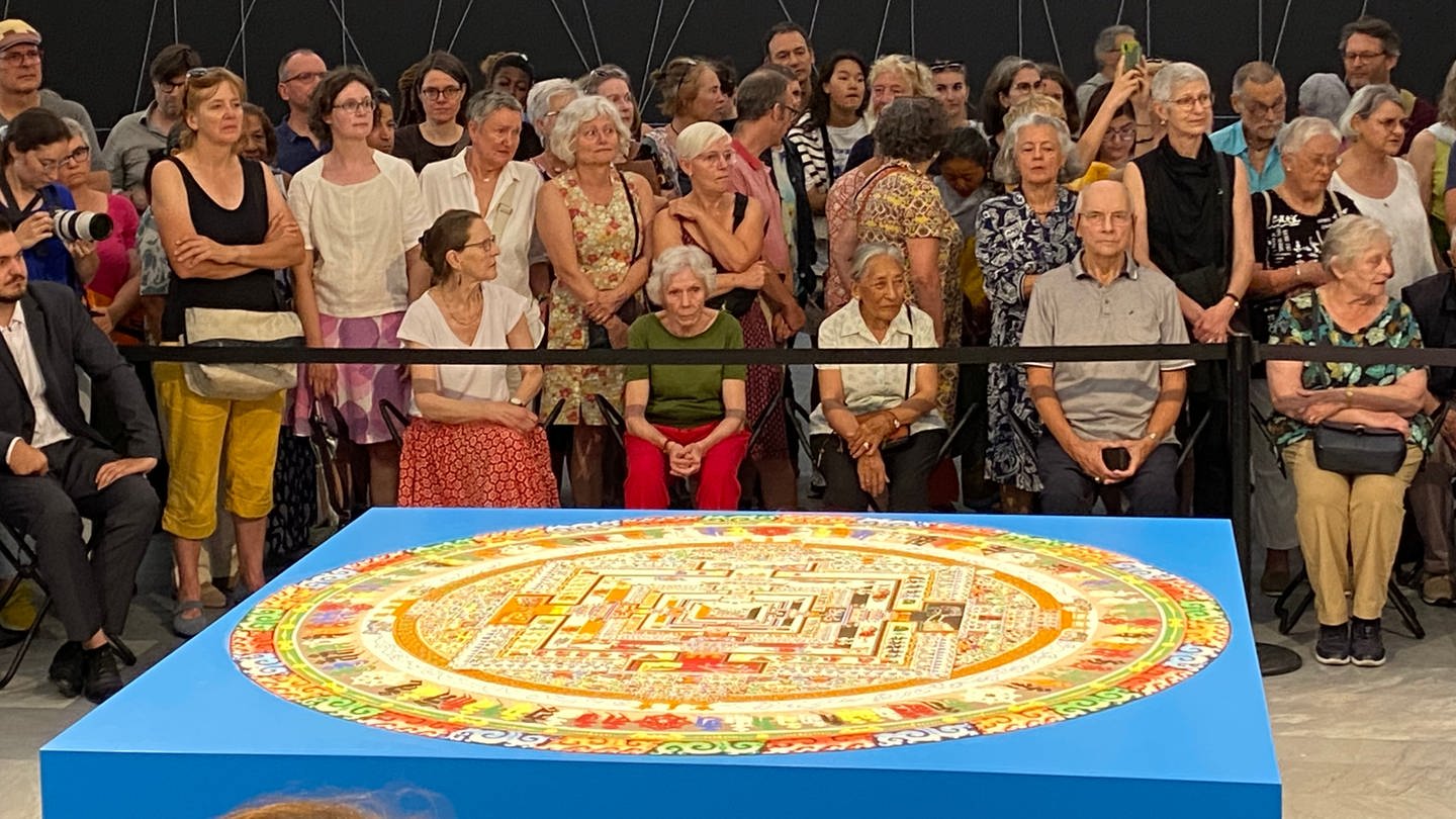 Zu sehen ist das bunte Kalachakra Mandala im Kunstmuseum Basel. Das Mandala ist rund und liegt auf einem blauen Würfel. Dahinter sitzt Publikum.