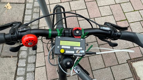 An einem Fahrradlenker ist ein rechteckiges Messgerät angebracht. Es soll den Abstand von Autos messen, die das Fahrrad überholen.