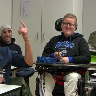 Junge mit blonden Haaren und Brille sitzt in einem selbstfahrenden Rollstuhl. Neben ihm ein Mann in Jacke. 