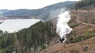 Schwarze Dampflok fährt an einem großen See vorbei. Über ihr Qualm.