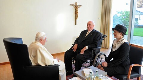 Papst sitzt gegenüber von einem älteren Mann mit weißen Haaren und Anzug. Neben ihm sitzt eine Frau mit Hut. 