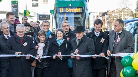 Mehrere Menschen stehen in einer Reihe, halten Scheren in der Hand und zerschneiden ein dünnes Band. Hinter ihnen steht eine grüne Straßenbahn mit der Aufschrift "8 Extrafahrt". 