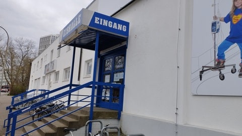 Zu sehen ist der Eingang der Echte Helden Arena in Freiburg. Die Wände der Arena sind weiß, das Treppengeländer zum Eingang ist blau. Der Eingang ist von der Seite fotografiert.