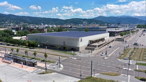 Von oben ist ein viereckiges, weißes Stadion zu sehen. Es hat ein blaues Dach mit Solapaneelen. Es liegt an einer Kreuzung in Freiburg. Dahinter ist die Stadt zu sehen, sowie der Schwarzwald. Der Himmel ist blau, es scheint die Sonne.