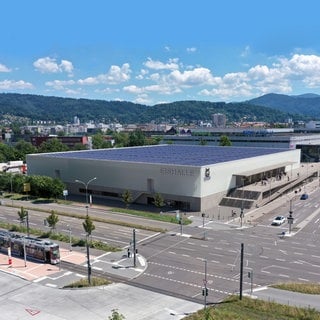 Von oben ist ein viereckiges, weißes Stadion zu sehen. Es hat ein blaues Dach mit Solapaneelen. Es liegt an einer Kreuzung in Freiburg. Dahinter ist die Stadt zu sehen, sowie der Schwarzwald. Der Himmel ist blau, es scheint die Sonne.