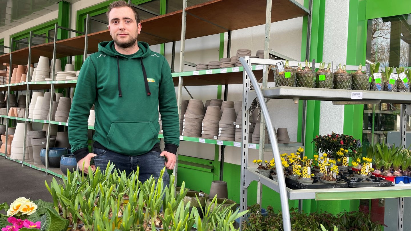 Ein Mann in grünem Pullover steht hinter einer Auslage mit Pflanzen. Hinter ihm sind Pflanzentöpfe zu sehen.