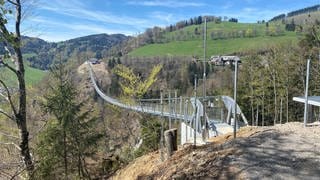 Eine silberene Hängebrücke aus Edelstahl überspannt ein Tal mit Wasserfall.