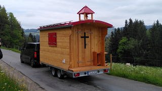 Schäferwagenkirche aus Holz wird als Anhänger von einem schwarzen Auto gezogen