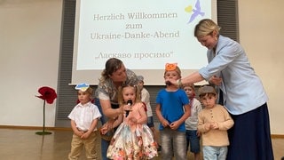 Zu sehen sind vier Kinder und zwei Frauen auf einer Bühne. Die Frauen halten den Kindern Mikrofone hin.