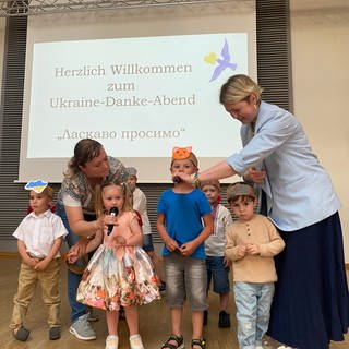 Zu sehen sind vier Kinder und zwei Frauen auf einer Bühne. Die Frauen halten den Kindern Mikrofone hin.