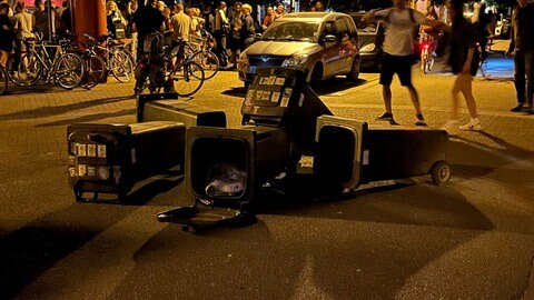 Mülltonnen liegen auf der Straße, im Hintergrund sieht man Menschen, die ausgelassen auf dem Lederleplatz in Freiburg feiern. 