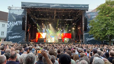 Auf dem Marktplatz in Lörrach findet ein Konzert vom Stimmen Festival statt. Im Hintergrund ist die Bühne zu sehen. Im Vordergrund des Bildes ist das Publikum zu sehen. Es ist von hinten fotografiert.