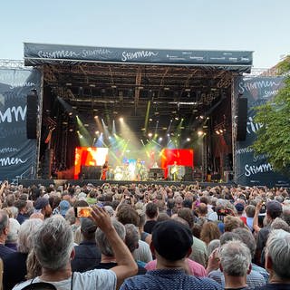 Auf dem Marktplatz in Lörrach findet ein Konzert vom Stimmen Festival statt. Im Hintergrund ist die Bühne zu sehen. Im Vordergrund des Bildes ist das Publikum zu sehen. Es ist von hinten fotografiert.