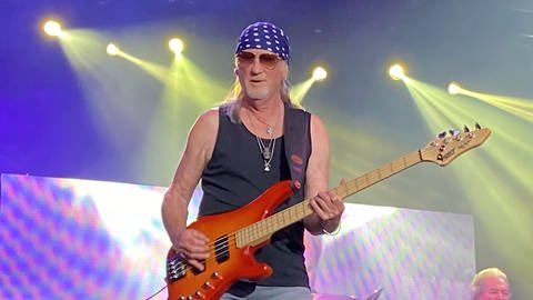 Deep Purple hat 2022 beim Stimmen-Festival in Lörrach gespielt. Zu sehen ist Roger Glover, der Bass spielt. Er trägt ein blaues Tuch auf dem Kopf mit weißen Punkten. Außerdem trägt er eine Sonnenbrille. Roger Glover trägt ein schwarzes Tanktop und eine helle Jeans.