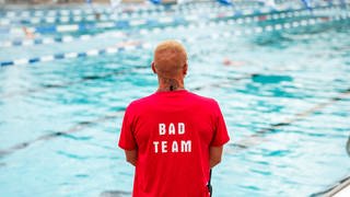 Ein Mann im roten T-shirt steht am Beckenrand in einem Freibad. Er ist von hinten zu sehen. Auf dem T-Shirt steht "Team Bad". Er hat blonde kurze Haare.
