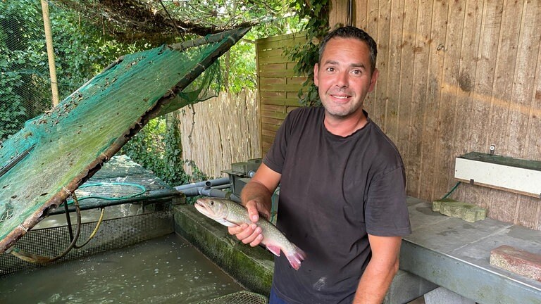 Ein Mann mit braunen, kurzen Haaren hält einen Fisch in der Hand. Er steht vor einem Wasserbecken. Der Mann ist ein Forellzüchter.