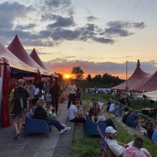 Auf dem Bild sieht man einen Sonnenuntergang im Hintergrund. Im Vordergrund sitzen Menschen auf Stühlen und einer Treppe zwischen Zelten und unterhalten sich.