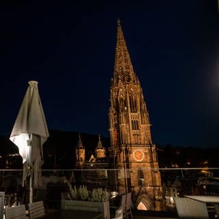Das beleuchtete Freiburger Münster bei Nacht, im Vordergrund eine Terrasse mit eingeklappten Schirmen.