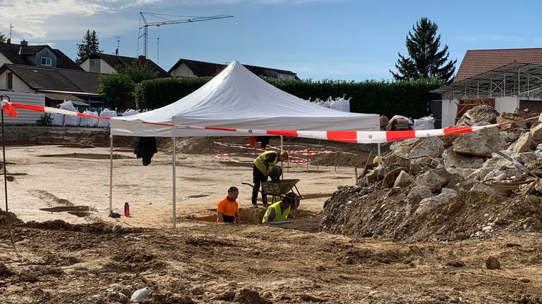 In der Mitte einer Baustelle steht ein Zelt, darunter graben Menschen in Warnwesten im Boden.