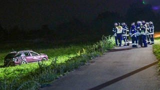 Feuerwehrleute und Polizisten stehen neben dem Wrack eines Autos, das zerbeult auf einer Weise neben der Straße steht.