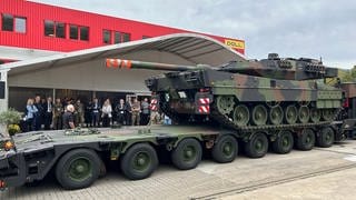 Ein Panzer steht auf einem Sattelanhänger. Im Hintegrund stehen Vertreter der Bundeswehr und Politik.