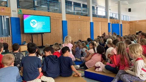 Grundschulkinder sitzen in der Turnhalle auf dem Boden. Sie schauen auf einen Bildschirm, auf dem die Verleihung des deutschen Schulpreises 2023 zu sehen ist.