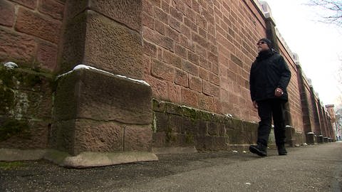 Ein Mann in schwarzer Kleidung und Sonnebrille läuft an der Gefängnismauer der Freiburger JVA entlang. 