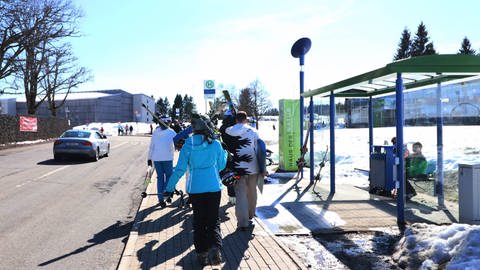 Ein paar Jugendliche laufen mit ihren Ski an einer Bushaltestelle auf dem Feldberg vorbei.