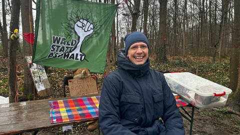 Mann mit kleiner runder Brille sitzt auf einer Bierzeltgarnitur, hinter ihm Plakate. Eins mit der Aufschrift "Wald statt Asphalt"