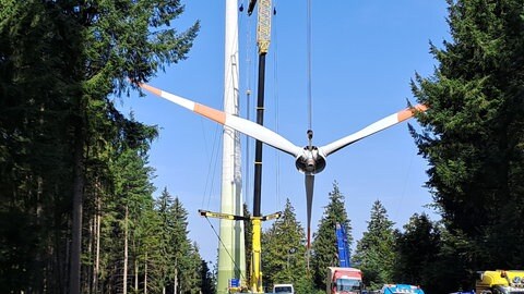 Die abmontierten Rotorenblätter eines Windrads hängen an einem Kran vor dem Betonturm. Unten sind Baufahrzeuge und Lastwagen zu sehen.