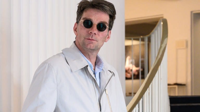 Mann mit runder Sonnenbrille und kurzen braunen Haaren schaut in die Kamera. Er trägt einen hellen Mantel und lehnt sich an ein Treppengeländer im Stil der 50er Jahre.