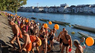 Zu sehen ist der Rhein in Basel. Im Rhein schwimmen viele Menschen. Am Ufer stehen noch mehr Menschen in Badekleidung, einige haben orangefarbene Luftballons in der Hand. Auf der anderen Seite des Rheins sind Häuser zu sehen.