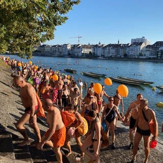 Zu sehen ist der Rhein in Basel. Im Rhein schwimmen viele Menschen. Am Ufer stehen noch mehr Menschen in Badekleidung, einige haben orangefarbene Luftballons in der Hand. Auf der anderen Seite des Rheins sind Häuser zu sehen.