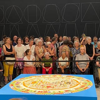 Zu sehen ist das bunte Kalachakra Mandala im Kunstmuseum Basel. Das Mandala ist rund und liegt auf einem blauen Würfel. Dahinter sitzt Publikum.