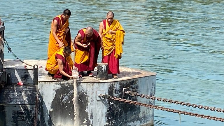Vier tibetische Mönche in orange-roten Gewändern stehen auf einer Plattform im Rhein. Einer der Mönche hält die Glasvase mit dem Sand in der Hand und schüttet den Sand in den Rhein. Hinter den Mönchen ist der Rhein zu sehen.