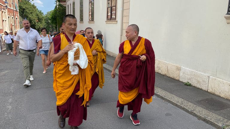 Zu sehen vorne zwei tibetische Mönche in rot-orangenen Gewändern. Der linke Mönch trägt eine Glasvase. In dieser befindet sich der Sand des Manalas. Dahinter läuft ein weitere Mönch und Menschen, die bei der Prozession zu sehen.