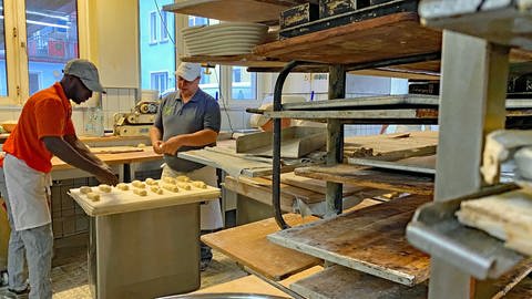 Die Bäckerei in der Abdoulie arbeitet ist zu sehen. Er rollt Teig und man sieht einige Regale für die Backwaren.