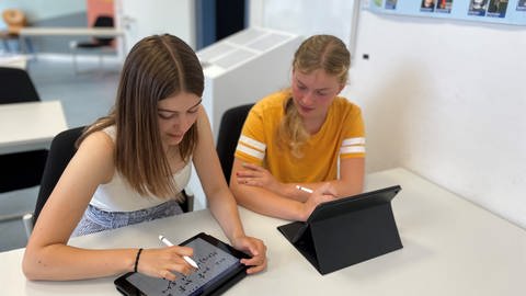 Die Schülerinnen Tabea und Friederike sitzen an einem Schreibtisch in einem Klassenzimmer.