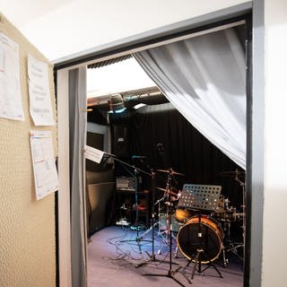 Hinter einer geöffneten Tür befindet sich ein leerer Proberaum. Darin steht unter anderem ein Schlagzeug.