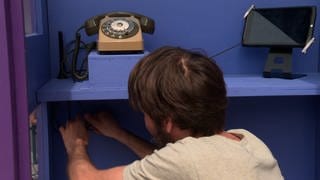 Ein Mann kniet vor der neuen Telefonzelle. Über ihm stehen ein altes Drehscheiben-Telefon und ein Tablet. 