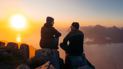 Ein Paar sitzt im Gegenlicht auf einer Bergkuppe, im Hintergrund ist das Meer zu sehen.