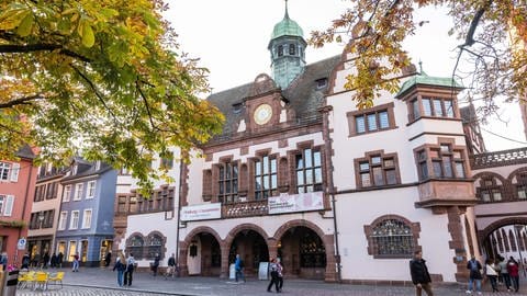 Herbstlich gefärbtes Laub hängt an den Bäumen auf dem Rathausplatz während dahinter das Freiburger Rathaus zu sehen ist.