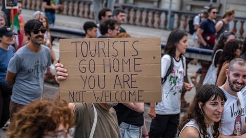 Ein Tourist hält bei einem Protestzug durch die Stadt ein Plakat in die Luft, auf dem er Touristen auffordert, wieder zu gehen.