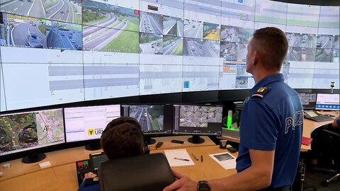 Ein Polizist steht vor einer Wand mit vielen Bildschirmen, auf dem der Verkehr auf den Autobahnen angezeigt wird.