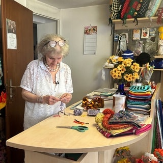 Gisela Mahler häkelt an der Ladentheke, im Hintergrund sind verschiedene Produkte ihrer Strick-Boutique in Bonndorf.