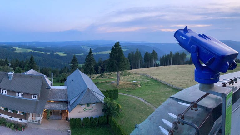 Von einer Aussichtsplattform, mit blauem Fernglas, sieht man den Feldberg und wie die Sonne langsam untergeht. Im Vordergrund befindet sich ein Hotelgebäude.